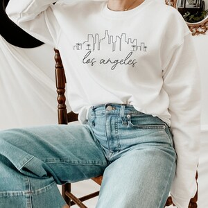 May include: A white sweatshirt with a black outline of the Los Angeles skyline and the text "los angeles" in cursive.