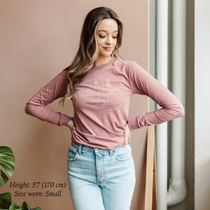 May include: A woman wearing a long-sleeved, mauve t-shirt with a white outline of the Raleigh, North Carolina skyline and the text "Raleigh, North Carolina". She is wearing light blue jeans and has long brown hair.