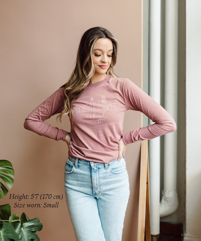 May include: A woman wearing a long-sleeved, mauve t-shirt with a white outline of the Raleigh, North Carolina skyline and the text "Raleigh, North Carolina". She is wearing light blue jeans and has long brown hair.