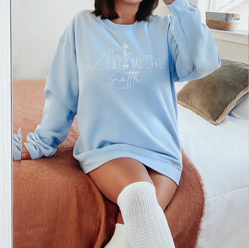 Pode incluir: Uma camisa de frio azul claro com um contorno branco do horizonte de Seattle e o texto "Seattle, Washington" na frente.