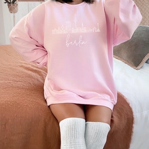 May include: A pink sweatshirt with a white outline of the Berlin skyline and the word "Berlin" written in cursive below the skyline.