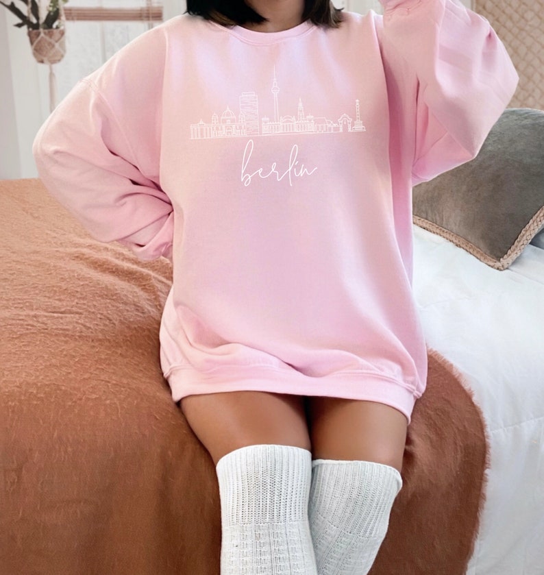May include: A pink sweatshirt with a white outline of the Berlin skyline and the word "Berlin" written in cursive below the skyline.