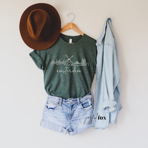 May include: A green t-shirt featuring a white outline of the Amsterdam skyline, including a windmill, and the word "amsterdam" written beneath. The t-shirt is hanging on a wooden hanger and is styled with denim shorts and a light blue denim jacket.