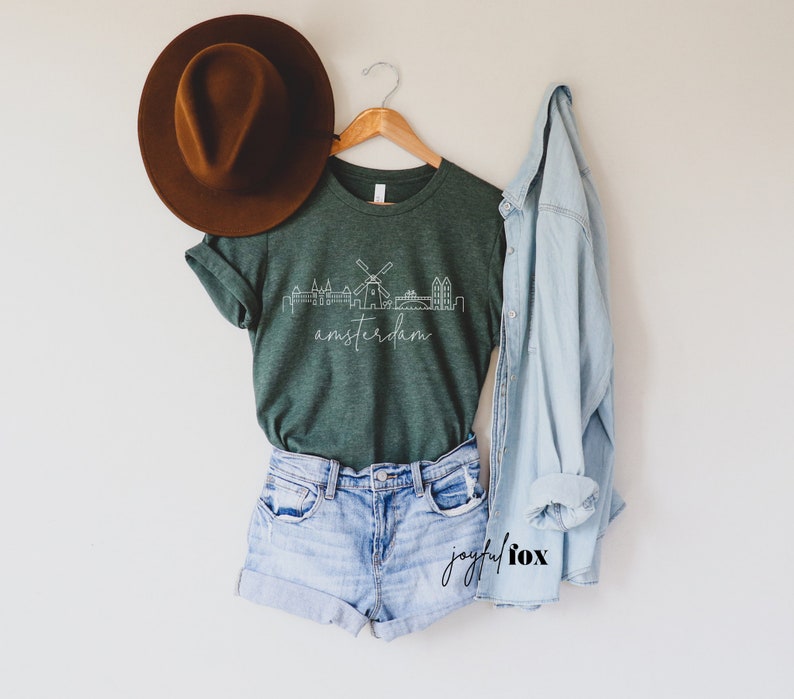 May include: A green t-shirt featuring a white outline of the Amsterdam skyline, including a windmill, and the word "amsterdam" written beneath. The t-shirt is hanging on a wooden hanger and is styled with denim shorts and a light blue denim jacket.
