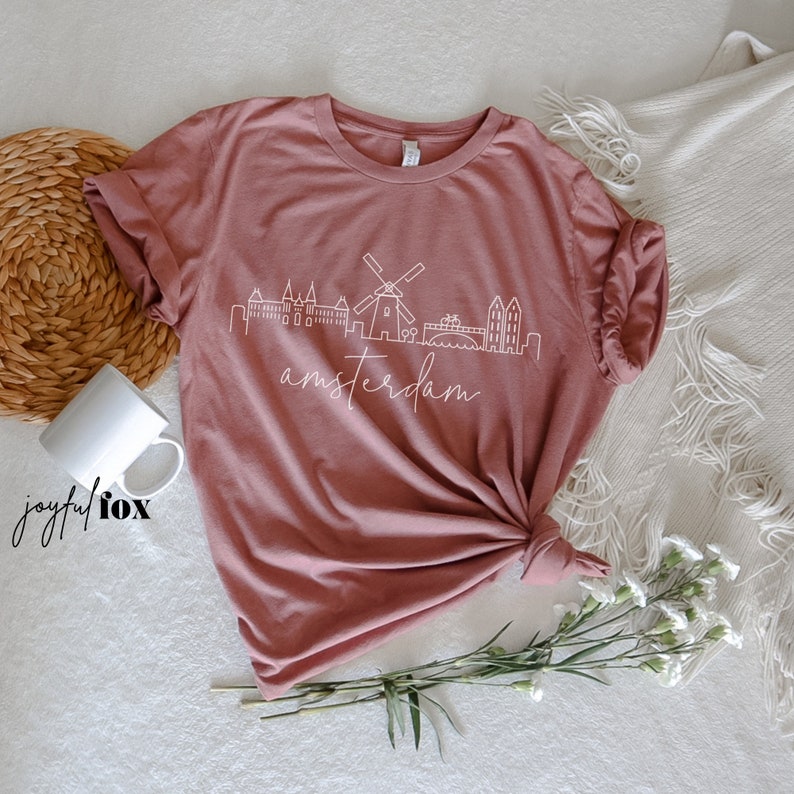 May include: A mauve t-shirt featuring a white outline of the Amsterdam skyline, including a windmill, with the word "amsterdam" written in cursive beneath.