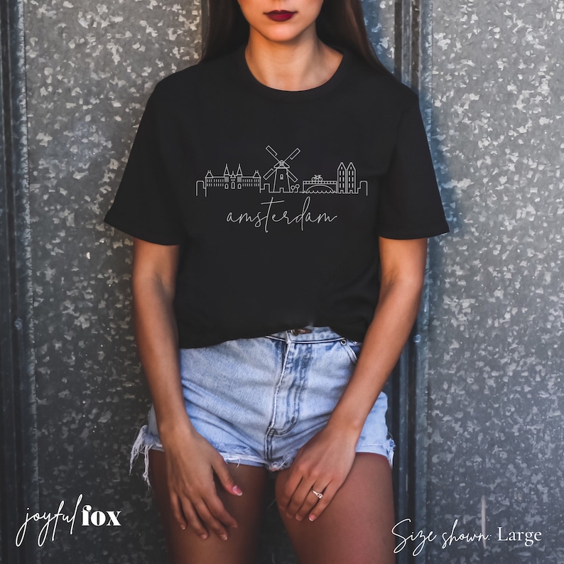 May include: Black t-shirt featuring a white outline of the Amsterdam skyline, including a windmill, and the word "amsterdam" written below the skyline. The size shown is large.