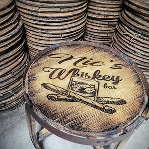 May include: A wooden barrel top stool with a burnt-in design that says "Mic's Whiskey Bar" with a glass of whiskey and two cigars. The stool is made from reclaimed wood and has a rustic look.