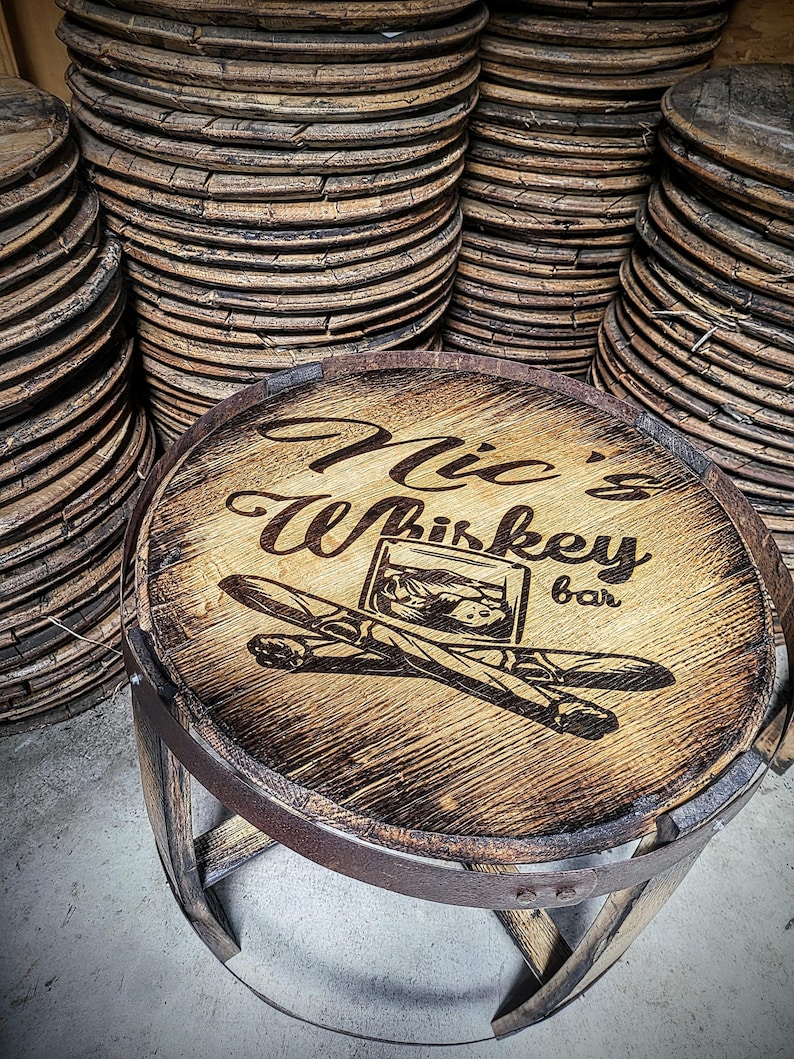 May include: A wooden barrel top stool with a burnt-in design that says "Mic's Whiskey Bar" with a glass of whiskey and two cigars. The stool is made from reclaimed wood and has a rustic look.