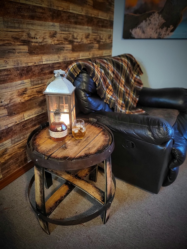 May include: A wooden barrel end table with a distressed finish, featuring a glass of whisky and a bottle of bourbon. The table is next to a black leather recliner with a plaid throw blanket.