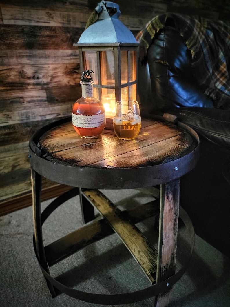 May include: A wooden barrel end table with a metal band around the top. A glass of amber liquid sits on the table next to a bottle of whiskey. A lantern with a candle inside is in the background.