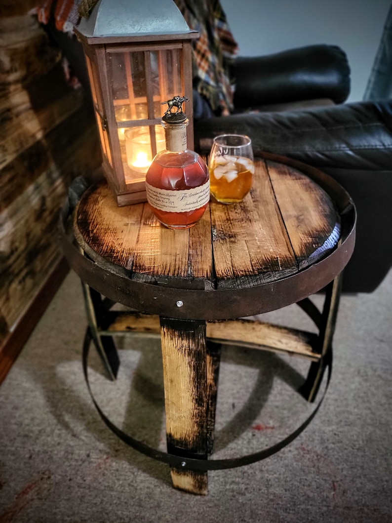 May include: A wooden barrel end table with a dark brown finish and black metal accents. A bottle of bourbon whiskey and a glass of iced tea are on the table.