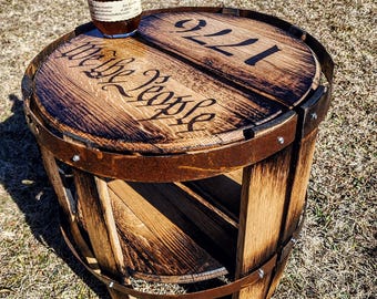 May include: A round wooden barrel table featuring a dark stained finish with the text "We the People" and "1776" burned into the top surface. The table includes a lower shelf and is secured with rusted metal bands. A bottle of Blanton's sits on top.