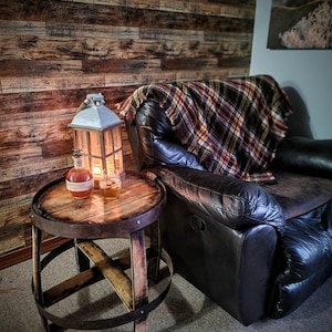 May include: A wooden barrel end table with a metal band around the top and bottom. The table has a distressed wood finish and is sitting on a carpet. A leather recliner with a plaid throw blanket is in the background. A lantern and a bottle are on the table.