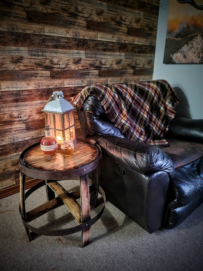 May include: A wooden barrel end table with a metal band around the top and bottom. The table has a distressed wood finish and is sitting on a carpet. A leather recliner with a plaid throw blanket is in the background. A lantern and a bottle are on the table.