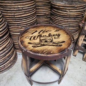 May include: A wooden bar stool with a circular seat featuring a wood-burned design that says "Nick's Whiskey Bar". The stool is made from reclaimed wood and has a rustic look.