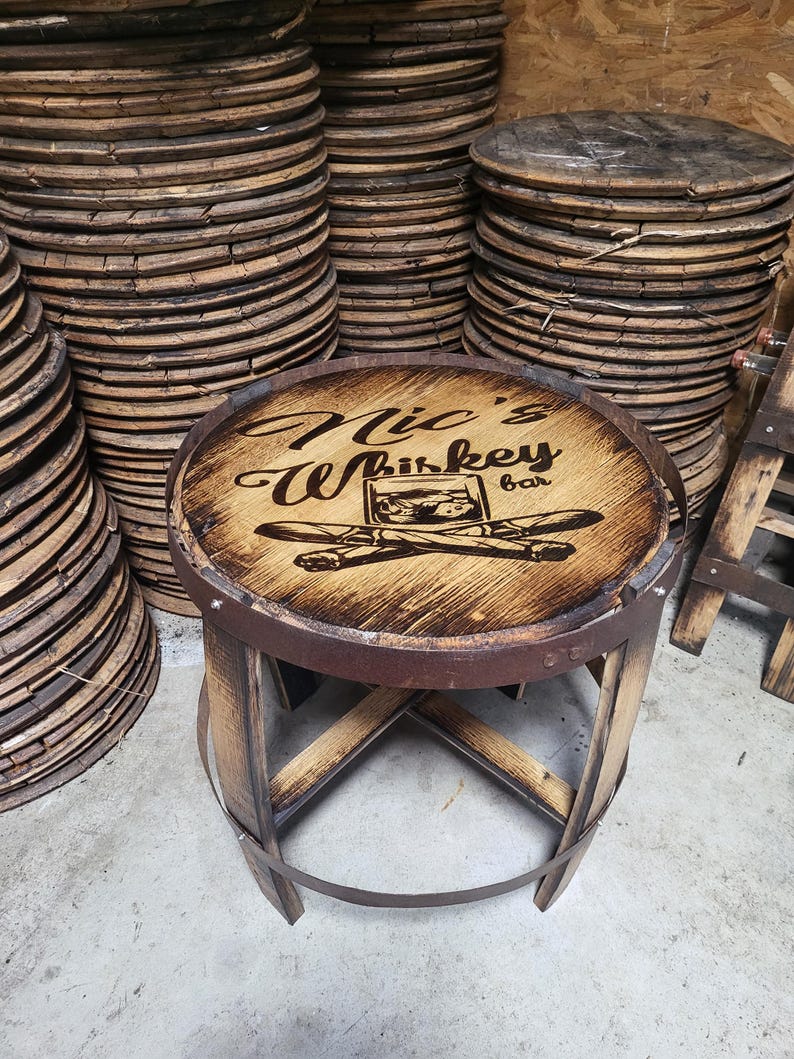 May include: A wooden bar stool with a circular seat featuring a wood-burned design that says "Nick's Whiskey Bar". The stool is made from reclaimed wood and has a rustic look.
