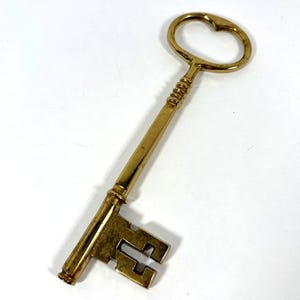 May include: A polished brass skeleton key featuring an oval-shaped bow, a slender cylindrical shank with a series of decorative ridges near the top, and a rectangular bit with a distinct cut-out notch pattern at the bottom end of the metal key.