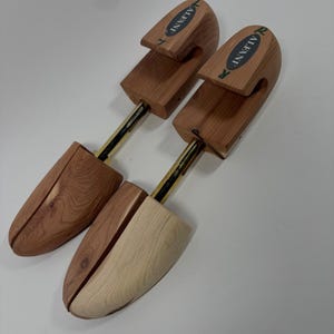 May include: A pair of wooden shoe trees with a natural grain finish and gold-toned metal spring mechanisms. Each heel piece features a green and white label with the text Alfani. The front sections are shaped to fit inside footwear.