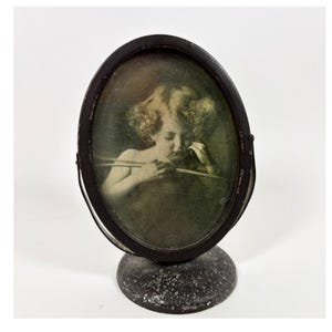 May include: An oval-shaped, dark metal tabletop frame featuring a vintage-style portrait of a child holding arrows. The frame is mounted on a circular, speckled grey base and includes a pivoting mechanism to adjust the angle of the portrait display.