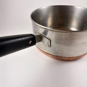 Könnte beinhalten: Ein Edelstahltopf mit kupferfarbenem Boden und einem langen, schwarzen Griff, der mit einer Metallhalterung und einer sichtbaren Schraube befestigt ist. Das Innere des Topfes ist glatt und metallisch und hat eine zylindrische Form mit flachem Boden.