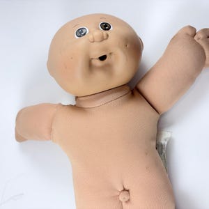 Puede incluir: Una muñeca con cabeza de vinilo lisa de tono claro, ojos marrones pintados, nariz pequeña y boca abierta. El cuerpo está confeccionado en tela suave de color beige claro con un ombligo visible y los brazos extendidos hacia afuera.