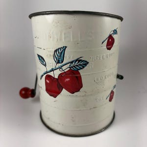 以下が含まれることがあります： 赤いクランクハンドルが付いた白い金属製の粉ふるい。外側には「Bromwell's」というエンボス加工の文字と、青い葉が付いた赤いリンゴのグラフィックがプリントされています。側面には1カップ、2カップ、3カップの目盛りが付いています。