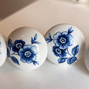 Op de afbeelding: Witte keramische kastknoppen met een blauw bloemmotief. De ronde knoppen hebben een gedetailleerd blauw bloemen- en bladerpatroon. Deze knoppen zijn waarschijnlijk voor een kast of lade.