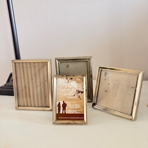 May include: Four rectangular photo frames with metallic gold-toned edges. The front-most frame contains a paper insert with the text 2.5 x 3.5 photo frame with non-glare glass, featuring a silhouette of two people and a sunset landscape.