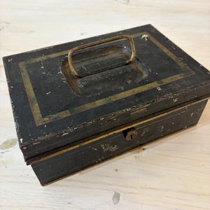 May include: A rectangular black metal cash box featuring a weathered finish with gold-toned border accents. The box includes a hinged metal handle on the top lid and a central keyhole on the front panel, showing signs of wear and paint chipping.