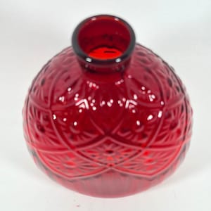 Puede incluir: Un jarrón de vidrio rojo intenso de forma redondeada, con un patrón geométrico en relieve en toda su superficie. El jarrón tiene una abertura circular estrecha en la parte superior y un cuerpo bulboso, mostrando un tono carmesí vibrante.