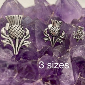 May include: Three silver thistle charms of different sizes on a bed of purple amethyst crystals. The text "3 sizes" is printed below the charms.