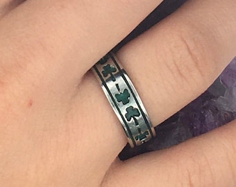 May include: A silver ring with a green clover design. The ring is worn on a person's finger.