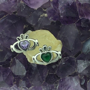 May include: Two silver Celtic knot earrings with heart-shaped stones. The left earring has a purple stone and the right earring has a green stone. The earrings are displayed on a bed of amethyst crystals.