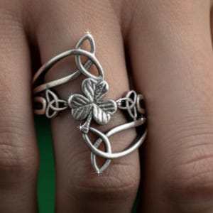 May include: A silver ring featuring a four-leaf clover design. The ring has intricate Celtic knot detailing and is worn on a finger. The clover is centered, with the knotwork extending around the band. The ring is made of a shiny metal.