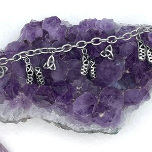 May include: A silver charm bracelet with multiple charms, including Celtic knots and a pair of dancing shoes. The bracelet is displayed on a bed of amethyst crystals.