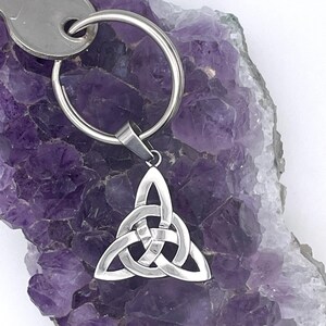 May include: A silver keychain featuring a Celtic knot pendant. The pendant, a three-pointed design, is attached to a key ring with a key. The background is a purple crystal.