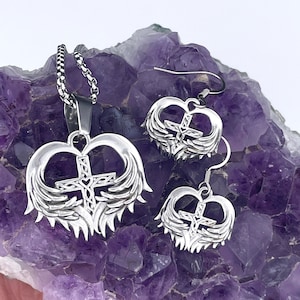 May include: A silver necklace and earrings set featuring a heart-shaped pendant with a cross and wings. The jewelry is displayed on a purple amethyst crystal.