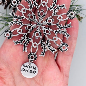Peut inclure: Ornement de Noël en forme de flocon de neige argenté avec un motif de chardon noir et une étiquette argentée qui dit "Nollaig Chridheil".