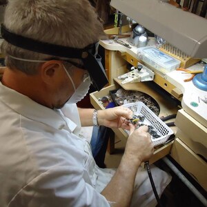 May include: A person wearing a white lab coat and a face mask is working on a small piece of jewelry. They are using a magnifying glass and tweezers to carefully examine the piece. The jewelry is in a small white basket on a workbench. There are various tools and supplies on the workbench, including a lamp, a pair of pliers, and a small container of beads.
