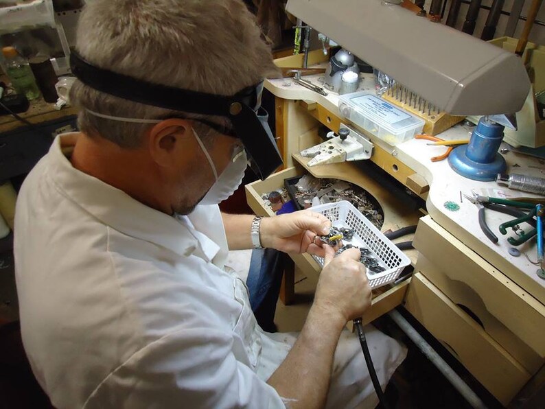 May include: A person wearing a white lab coat and a face mask is working on a small piece of jewelry. They are using a magnifying glass and tweezers to carefully examine the piece. The jewelry is in a small white basket on a workbench. There are various tools and supplies on the workbench, including a lamp, a pair of pliers, and a small container of beads.