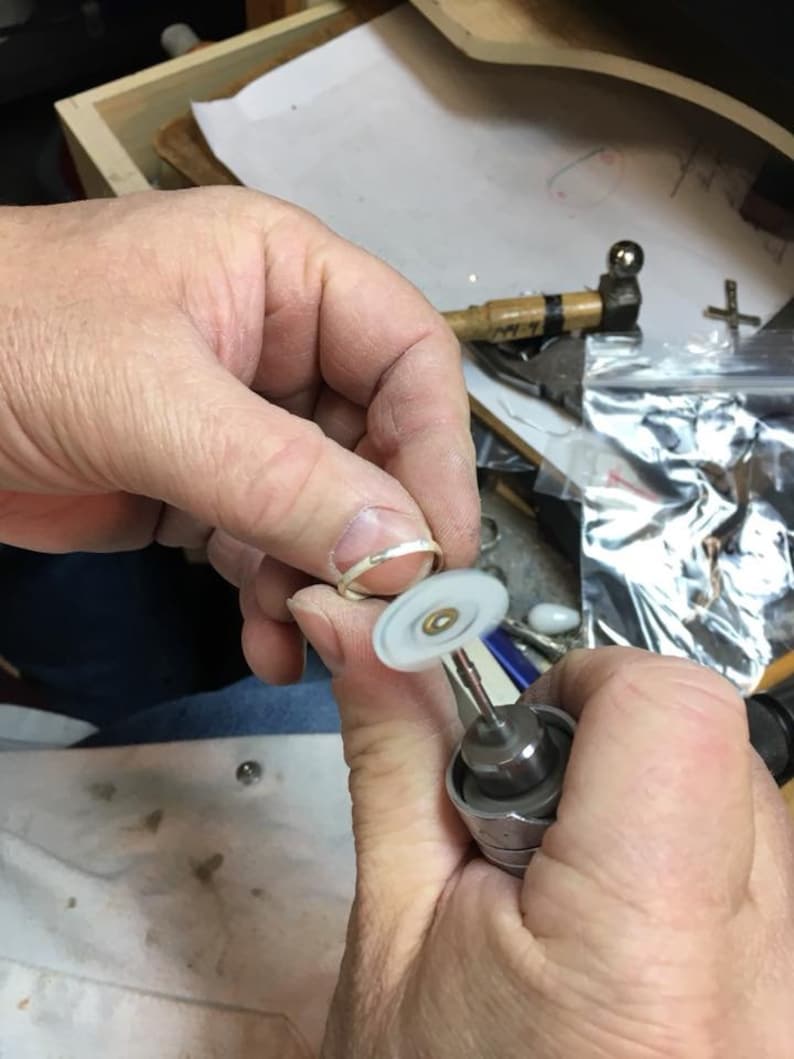 May include: A person is using a small, handheld rotary tool with a white polishing wheel to polish a silver ring.