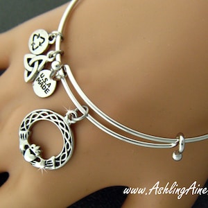 Könnte beinhalten: Silbernes Armreif-Armband mit drei Anhängern: einem keltischen Knoten, einem Kleeblatt und einem Kreis mit einem Claddagh-Design. Die Anhänger sind mit Sprungringen am Armband befestigt. Das Armband ist in den USA hergestellt.