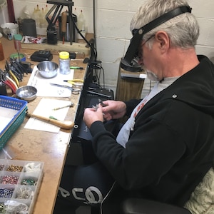 May include: A person wearing a black sweatshirt and a magnifying headset is working on a jewelry project at a workbench. The workbench is covered in tools and supplies, including a small container of beads, a pair of tweezers, and a small metal bowl.
