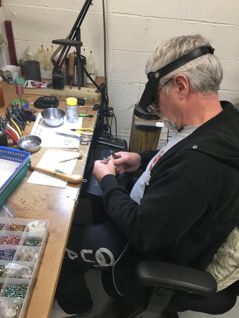 May include: A person wearing a black sweatshirt and a magnifying headset is working on a jewelry project at a workbench. The workbench is covered in tools and supplies, including a small container of beads, a pair of tweezers, and a small metal bowl.