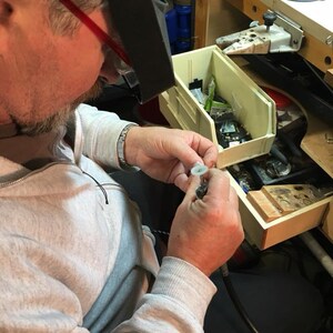 May include: A person wearing a grey sweatshirt and a magnifying visor is working on a small object with a tool. The person is sitting at a workbench with a variety of tools and supplies.