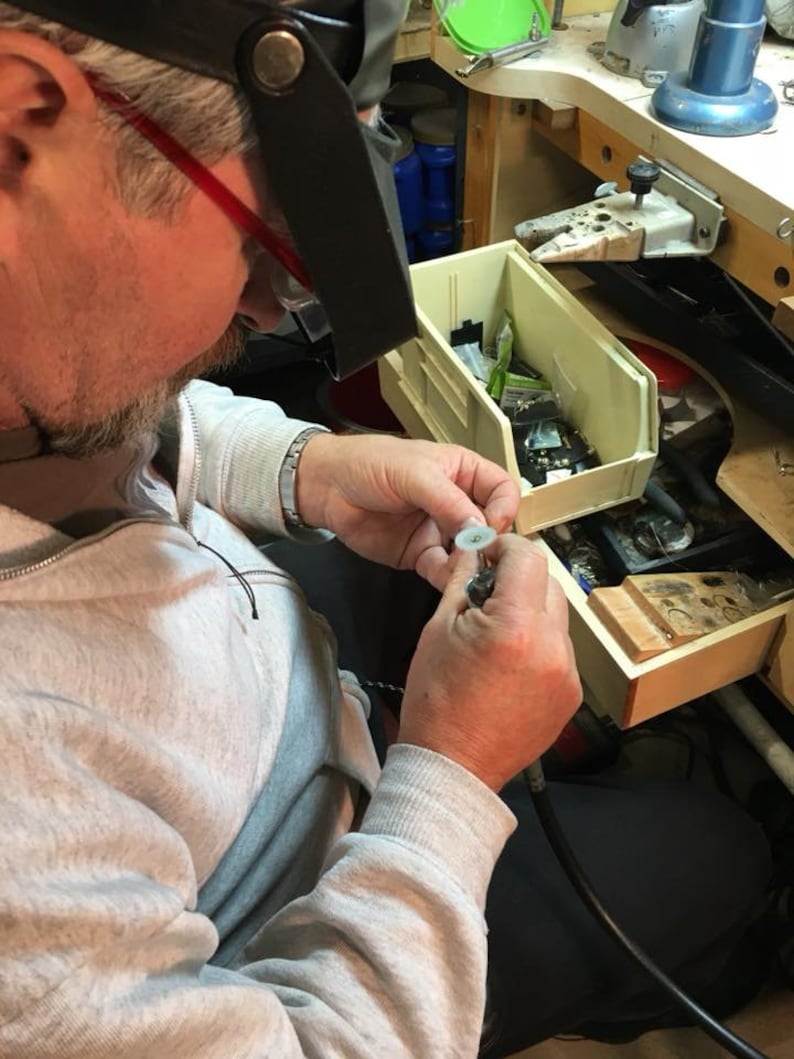 May include: A person wearing a grey sweatshirt and a magnifying visor is working on a small object with a tool. The person is sitting at a workbench with a variety of tools and supplies.