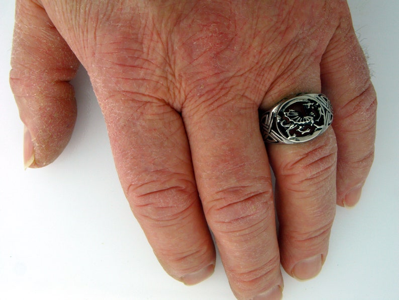 May include: A silver ring with a detailed design of a dragon on the top. The ring is worn on the middle finger of a hand.
