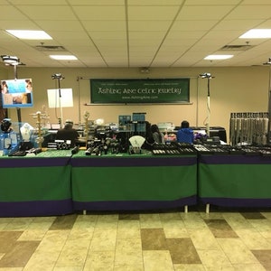 May include: A jewelry vendor's booth at a craft fair, featuring a variety of silver and gemstone jewelry. The booth is decorated with green and purple tablecloths and has a sign that reads "Ashing Ane Celtic Jewelry" and a website address.