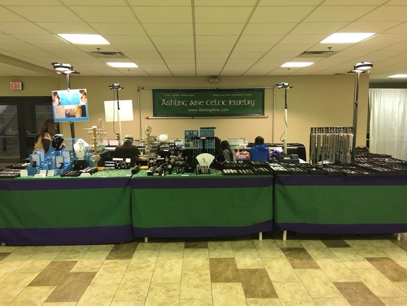 May include: A jewelry vendor's booth at a craft fair, featuring a variety of silver and gemstone jewelry. The booth is decorated with green and purple tablecloths and has a sign that reads "Ashing Ane Celtic Jewelry" and a website address.