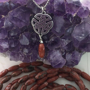 May include: A silver Celtic knot pendant with a purple enamel design, hanging from a silver chain. A red gemstone bead dangles from the pendant.